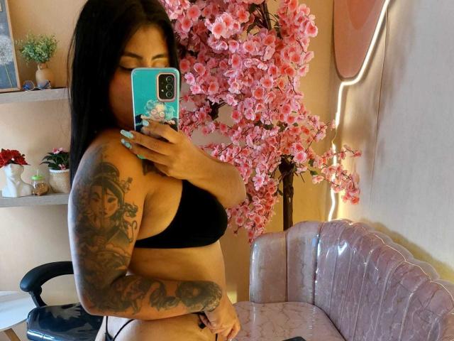 AngelaJoness with black hair, wearing a black bra, showing a tattooed arm, holding a phone with a pink floral case, standing in front of pink cherry blossoms.