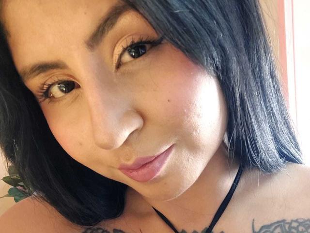 AngelaJoness has medium brown skin, black shoulder-length hair, brown eyes with long lashes, and a small tattoo on her left shoulder. Her lips are slightly puckered.