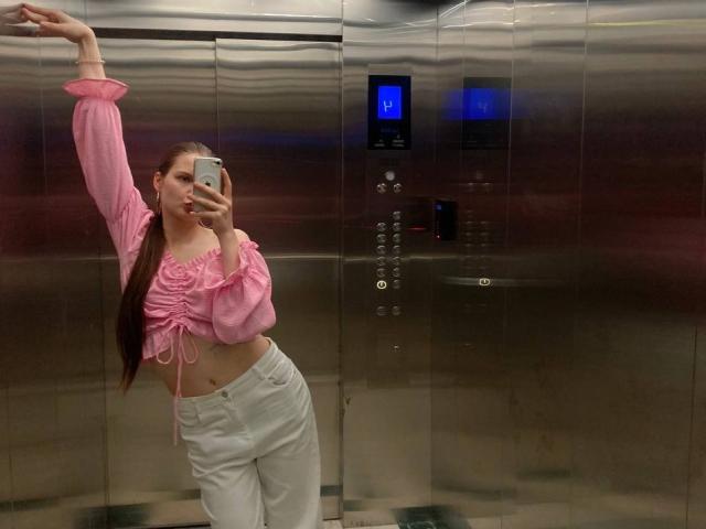 JuttaHillwig with long brown hair, pink off-shoulder top, white pants, arm raised, taking mirror selfie in elevator, showing midriff, small ass, slim figure, phone in hand.
