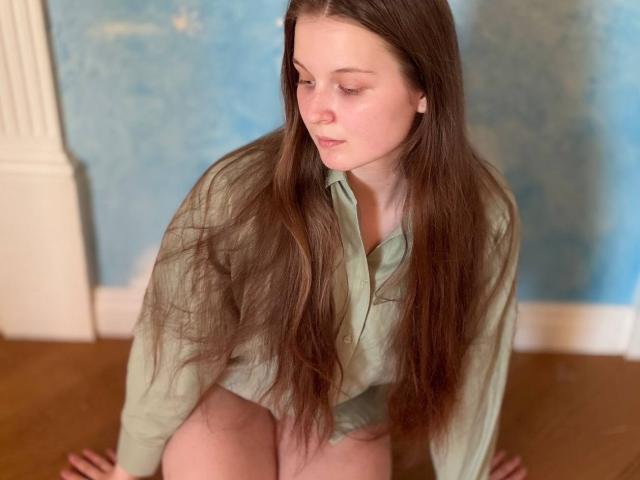JuttaHillwig with long brown hair, green shirt, and no pants, kneeling on wooden floor, looking down, hands on knees, soft pink lips, pale skin, slightly flushed, relaxed expression.