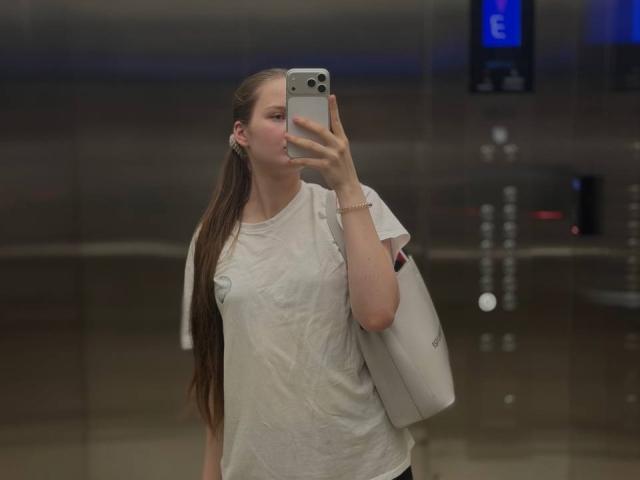 JuttaHillwig with long brown ponytail, wearing a wet white shirt, holding a phone in front of her face, white bag over shoulder, standing in a dimly lit elevator.