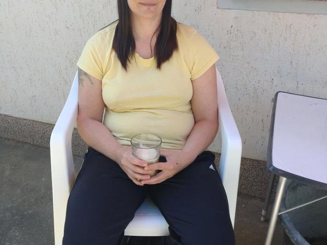 YviEngel87: una ragazza attraente con il seno scoperto, indossa una camicia gialla e pantaloni neri, tiene in mano un bicchiere con un liquido bianco, è seduta su una sedia bianca, con le braccia appoggiate sulle cosce, ha i capelli castani..