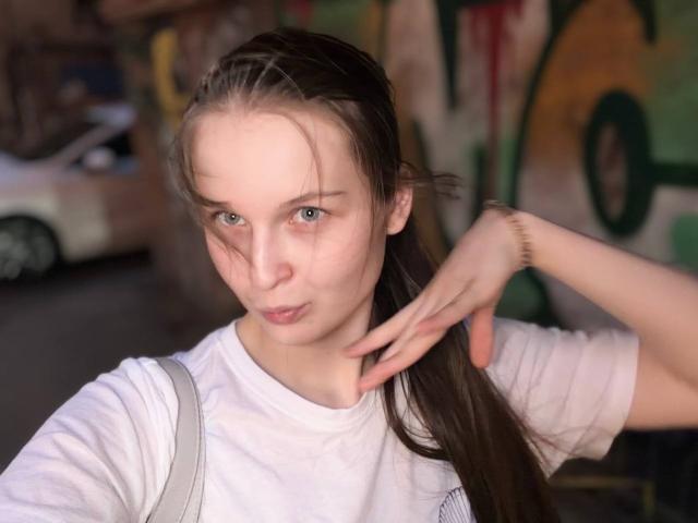 JuttaHillwig has pale skin, blue eyes, and long brown hair, wearing a white shirt, with a hand near her neck, looking slightly pouty. Background is blurred.
