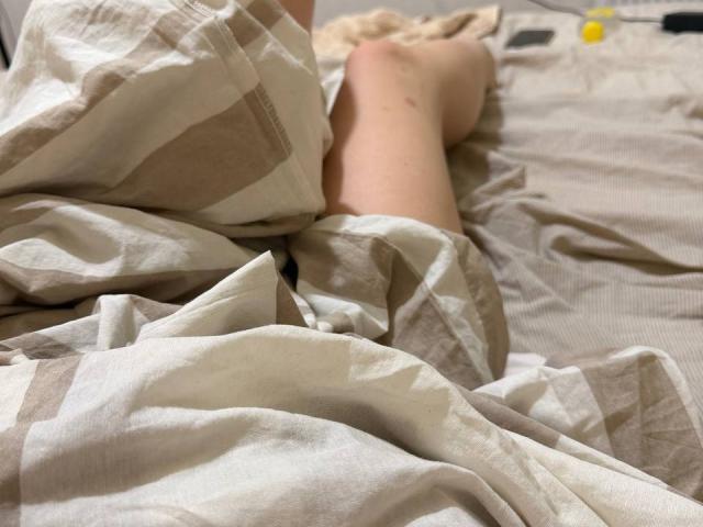 JuttaHillwig's legs are under a beige and white striped bedsheet, with her bare feet visible in the background. A yellow bottle and black object are on the bed.