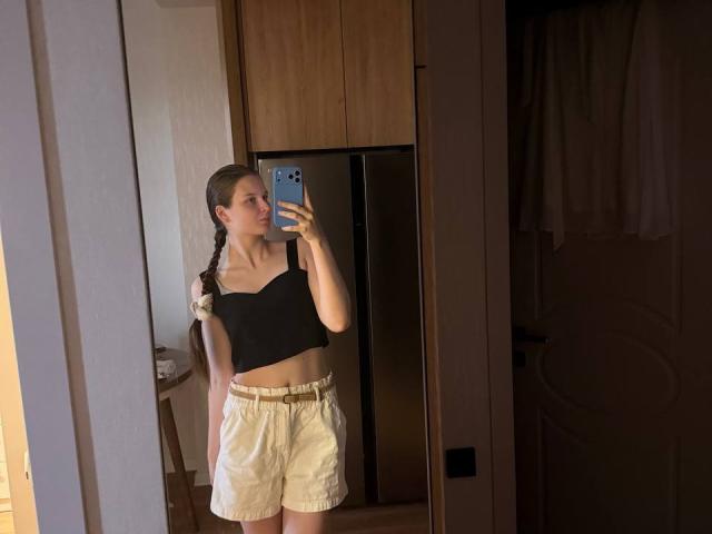 JuttaHillwig with long braids, black crop top, and yellow shorts, standing in a room, taking a mirror selfie with a blue phone, showing a slim, flat-ass.