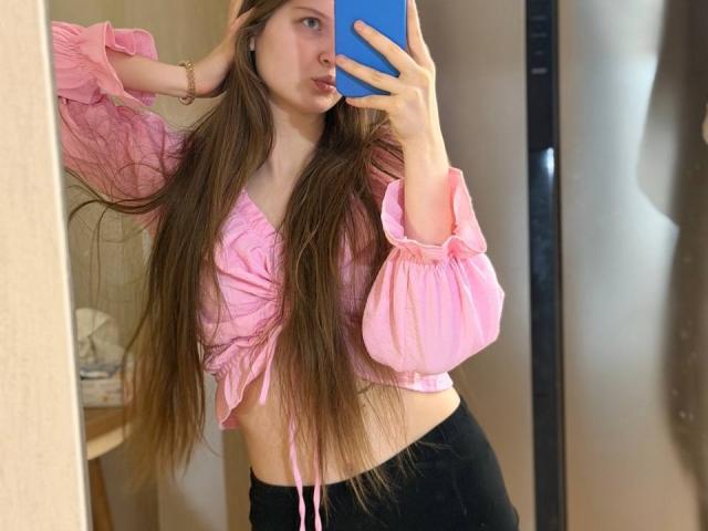 JuttaHillwig with long brown hair, pink ruffled crop top, black pants, holding blue phone, mirror selfie, showing her tits, cute, slim, fair skin, hand in hair.