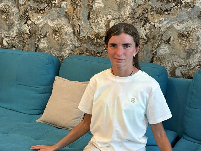 DarsiSweet with brown hair in a white t-shirt, sitting on a blue couch with a beige pillow, looking at the camera, relaxed. Rock-patterned wall in background.