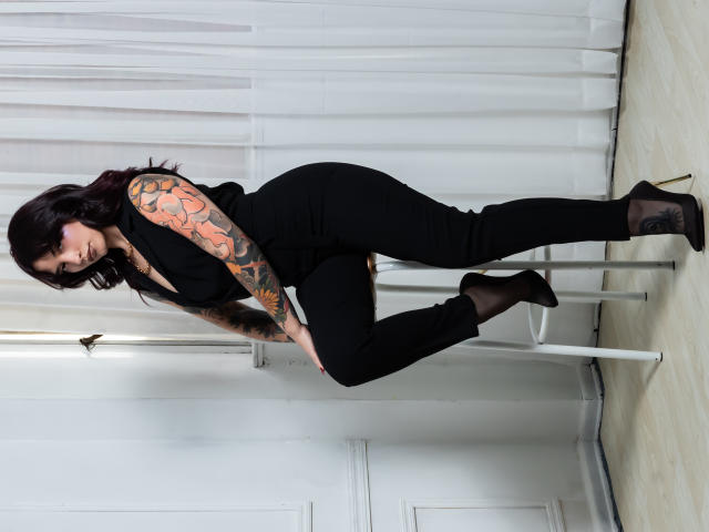 AkiraNishimura69, met een getatoeëerde arm, zwarte broek en hakken, leunt tegen een witte paal, en laat haar billen en borsten zien in een strak zwart outfit, met donker haar..