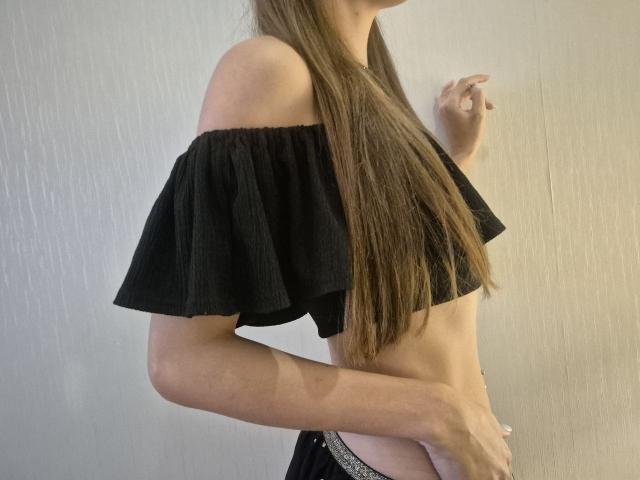 TenerliRize with long brown hair, black off-shoulder crop top, showing small tits, slim waist, pointing with right hand, wearing black pants with white lace trim.