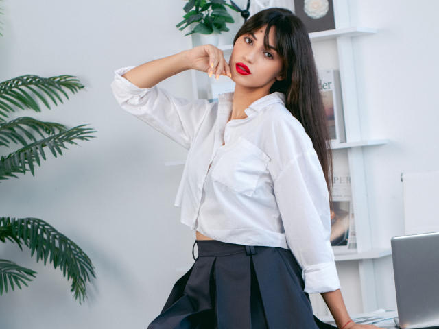 JasmineDay with dark hair, red lipstick, white shirt, and black skirt, standing with hand near mouth, looking at camera, in a bright room with plants and shelves.