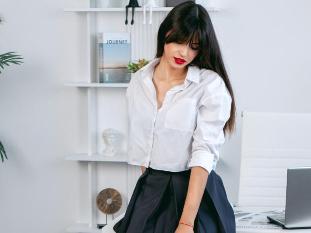 JasmineDay with long dark hair, red lipstick, white button-up shirt, and black skirt, adjusting her skirt, showing her smooth, curvy ass, standing in a bright office.