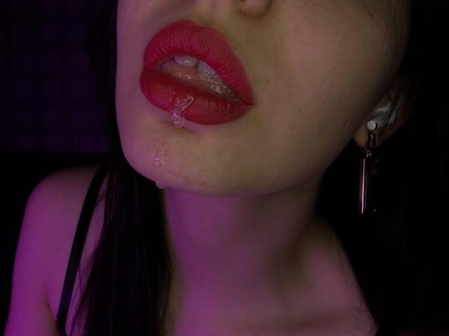 TracyVerbeke's glossy red lips are slightly parted with a droplet of saliva on her lower lip, wearing a black strap, and has a pinkish-purple light on her face.