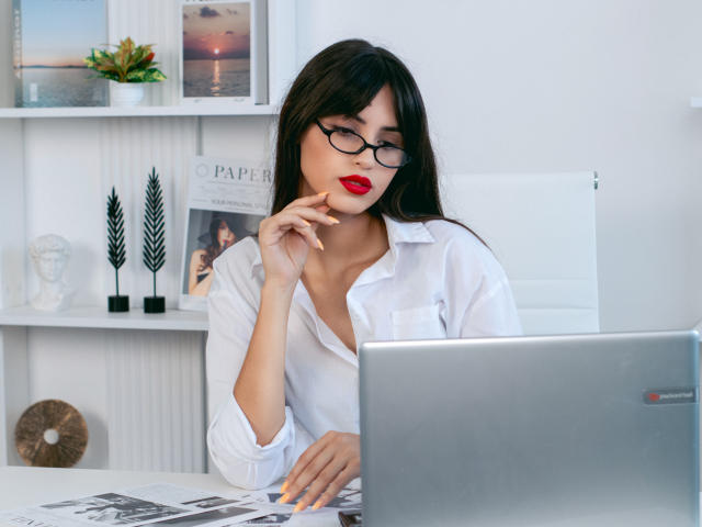 JasmineDay with black glasses, red lipstick, and white shirt, finger on chin, looking at laptop, sitting at desk with magazine, has long black hair, sexy.