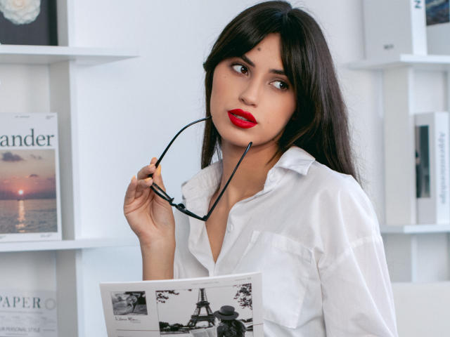 JasmineDay with long black hair, red lipstick, white shirt, holding black glasses, looking at a magazine with a black-and-white Eiffel Tower photo.