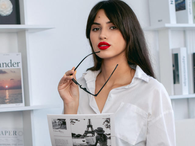 JasmineDay with dark hair, red lipstick, white button-up shirt, holding glasses in mouth, looking at black-and-white magazine, has medium tits, white shelves in background.