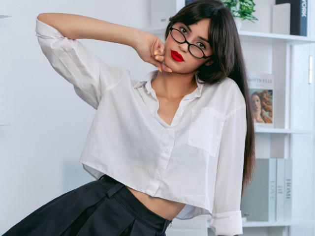 JasmineDay with long black hair, wearing a sheer white button-up shirt exposing her small tits and black skirt, leaning on a white shelf, wearing glasses and red lipstick.