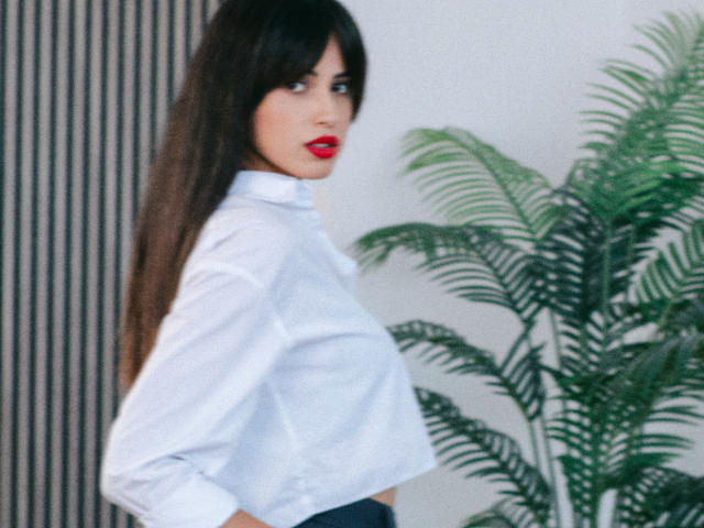 JasmineDay with long black hair, red lipstick, white shirt, and dark pants, standing sideways, showing her tits and looking over her shoulder. Potted green plant in background.