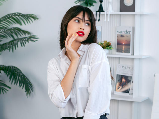 JasmineDay with red lipstick, white button-up shirt, dark hair, and hand on cheek, looking to the side, standing in front of a bookshelf with green plants.