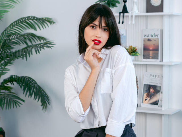 JasmineDay with dark hair, red lipstick, white shirt, black skirt, holding lip with finger, looking at camera, small ass, pensive expression, green plant on left.