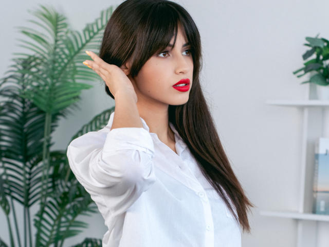 JasmineDay with long black hair, red lipstick, white button-up shirt, and raised hand, showing off her tits slightly. She's looking to the side with a sexy, confident look.