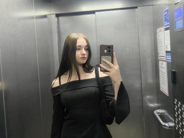 Lixsi mit langen braunen Haaren, schwarzem, schulterfreien Kleid, hält ein Telefon, Selfiemotiv im Aufzug, schwarze Tasche über der Schulter, graue, metallische Wände, neutraler Gesichtsausdruck, durchschnittlich große Brüste..