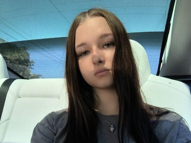 Lixsi a de longs cheveux bruns, une peau claire et une expression neutre. Elle porte une chemise grise avec un petit collier et est assise dans un siège de voiture blanc..