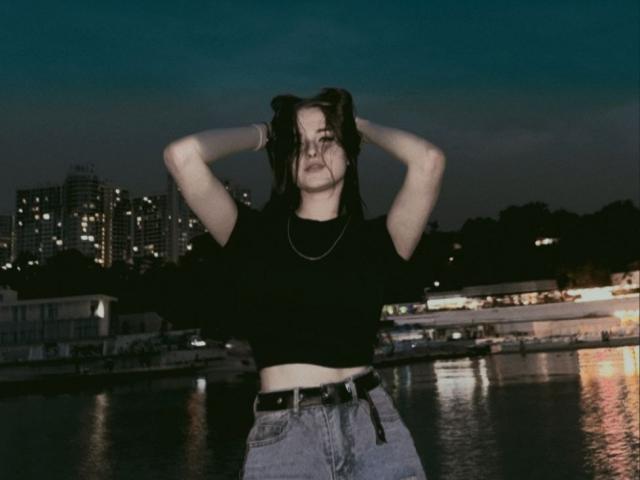 Lixsi hat ihre Hände in ihrem dunklen Haar vergraben, trägt ein schwarzes Crop-Top und hochgeschnittene Jeans und steht nachts an einem glitzernden Wasserlauf, im Hintergrund sind die Lichter der Stadt zu sehen..