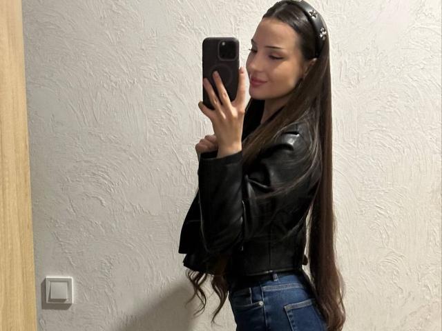 Versavi with long brown hair, black leather jacket, and blue jeans, taking a mirror selfie, holding a black phone, smiling, wearing a headband, white textured wall in background.