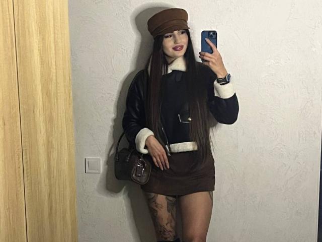 Versavi has long brown hair, wears a brown cap, black and white sweater, brown mini-skirt, and brown purse. She's taking a mirror selfie, showing a tattoo on her thigh.