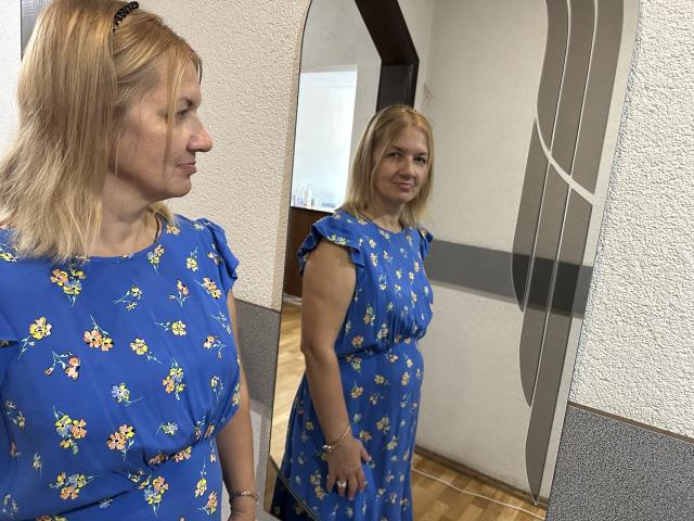 MelisaWorld, con cabello rubio, vestida con un vestido azul con estampado floral, está de pie frente a un espejo, mostrando su reflejo, con una sonrisa sutil, en una habitación con paredes texturizadas..