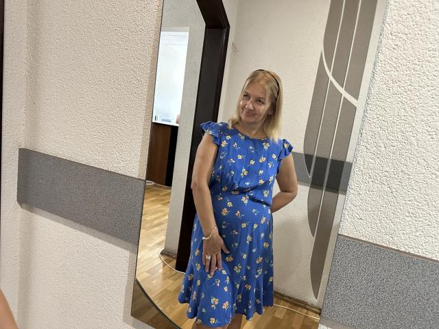 MelisaWorld staat lachend in een blauwe jurk met witte margrietmotieven, handen op haar heupen, blond haar, voor een spiegel, met een houten vloer, witte, gestructureerde muren en een grijze paneel..