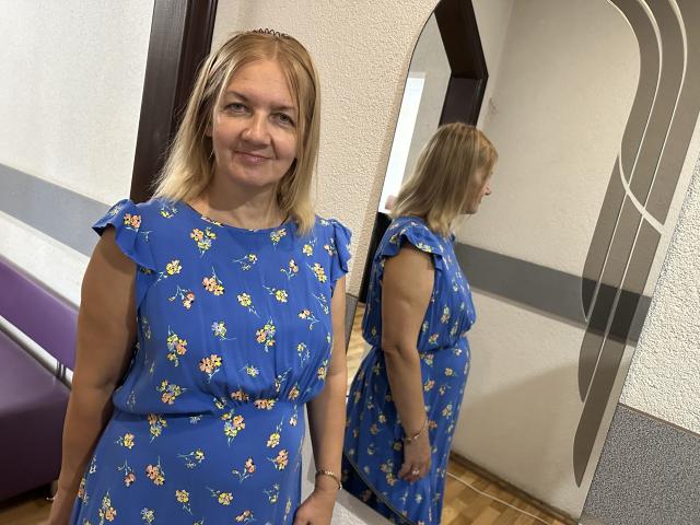 MelisaWorld, com cabelo loiro, sorrindo, vestindo um vestido azul com estampas florais e mangas bufantes, refletida em um espelho. Seus seios são discretos, e ela está em pé em um quarto..