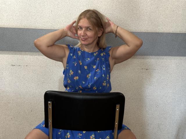MelisaWorld, blonde, muskulöse Arme, trägt ein blaues Kleid mit Blumenmuster, sitzt auf einem schwarzen Stuhl, Hände im Haar, lächelt, Po leicht sichtbar, Foto, lässig, attraktiv, selbstbewusst, entspannt..