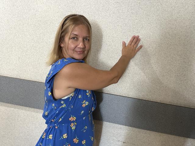 MelisaWorld, con i capelli biondi, indossa un abito blu con fiori e sta appoggiata a un muro, sorridendo. Una mano tocca il muro, mostrando leggermente il sedere. Il suo seno è piccolo..