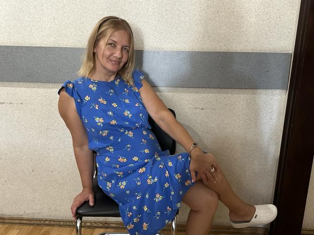 MelisaWorld hat blondes Haar, ein blaues Kleid mit Blumenmuster, weiße Turnschuhe und ein Armband. Sie sitzt auf einem schwarzen Stuhl in einem einfachen Raum, ein Bein übereinander geschlagen, und lächelt..
