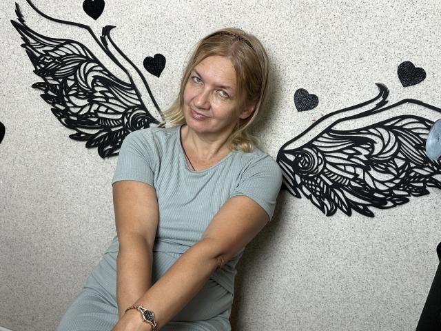 MelisaWorld, con i capelli biondi, la pelle chiara e il seno piccolo, indossa una maglietta grigia a coste e siede contro un muro con adesivi neri a forma di ali e cuore, sorridendo leggermente..