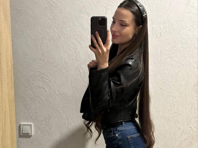 Versavi with long brown hair, black leather jacket, and blue jeans, taking a mirror selfie with a pouty expression, holding a black phone, wearing a black headband.