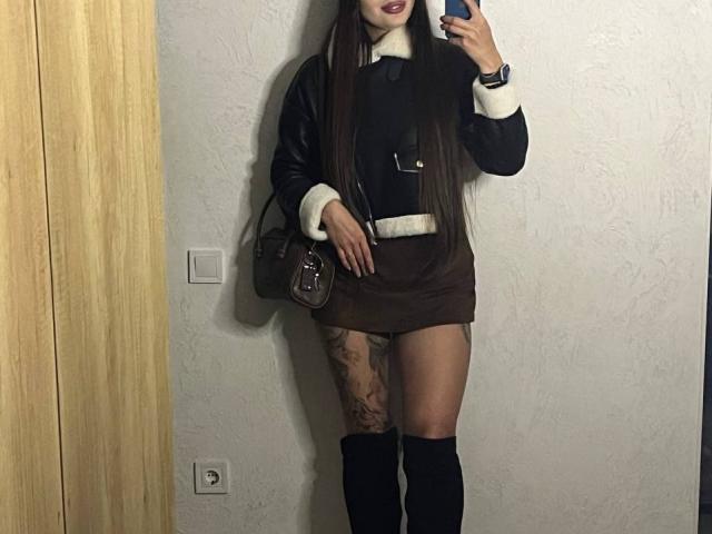 Versavi with long brown hair, wearing a black leather jacket, brown mini-skirt, white fur trim, black knee boots, holding a phone, and a brown purse, tattooed thigh visible.