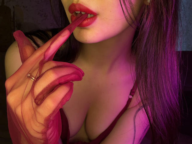 TracyVerbeke with red lipstick, biting her pink-red sheer fingerless glove, showing her tits, and wearing a ring on her finger. Her purple hair is in the background.