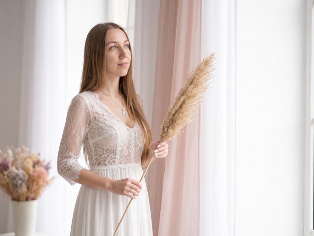EllaMint with long brown hair, wearing a white lace dress, holding a pampas grass, standing near a window, smiling, looking to the side, with a floral vase.