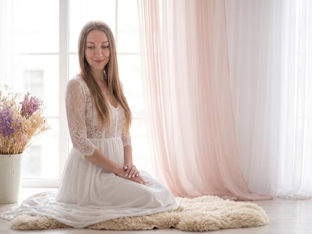 EllaMint, with long brown hair, sits on a fluffy rug, wearing a white lace dress, smiling gently, hands on knees, in a bright, softly lit room.