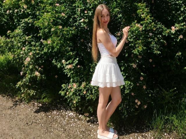 EllaMint with long blonde hair, white tank top, and white lace skirt, standing on a dirt path, holding pink roses, wearing white sandals, smiling, holding roses.