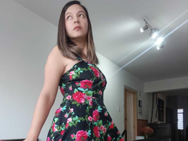 ArianaBlonde with medium brown hair, wearing a black floral dress with pink roses, has a curvy butt and small tits, standing in a room with a ceiling light.