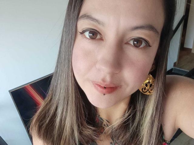 ArianaBlonde has light brown eyes, long brown hair with blonde highlights, a lip piercing, and is wearing ornate gold earrings, looking at the camera with a slight smile.