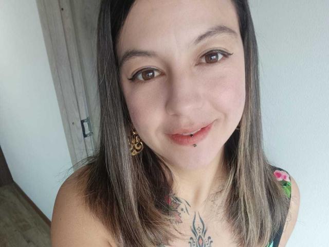 ArianaBlonde has light skin, brown eyes with winged eyeliner, a lip piercing, and a tattoo on her chest. She's smiling with straight brown hair, wearing a floral top.