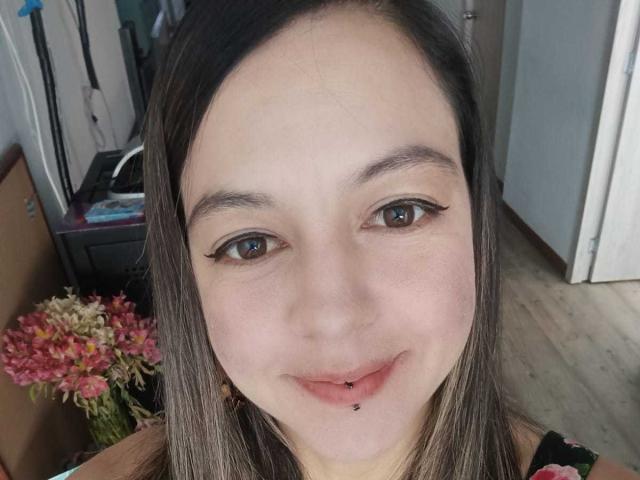 ArianaBlonde has fair skin, brown eyes with winged eyeliner, and a pierced lower lip. She's smiling with straight brown hair and wearing a floral top.