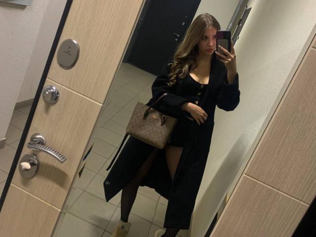 MarcelineConkey with long brown hair, black dress, thigh-high slit, black stockings, boots, and brown handbag, taking a mirror selfie in a bathroom, holding a phone.