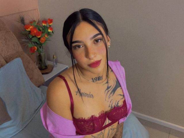 SophiieBlair with black hair, red lipstick, purple off-shoulder top, red lace bra, tattoos, and earrings, sitting on a bed with a bouquet of orange roses in the background.