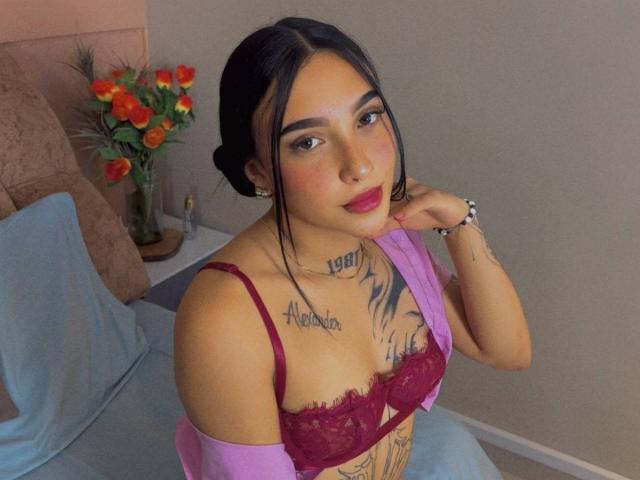 SophiieBlair with dark hair, pink lipstick, red bra showing her tits, tattoos on chest and neck, pink off-shoulder top, hand on cheek, flowers in background.