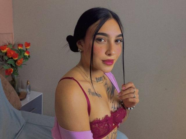 SophiieBlair with black hair in a bun, wearing a red lace bra, pink sleeves, holding a hairpin, tattoos on chest, pink lipstick, gray wall, vase with red roses.